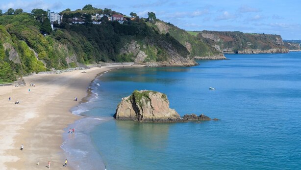 A coastal beach with a rocky outcrop, clear blue water, and houses on a hillside.