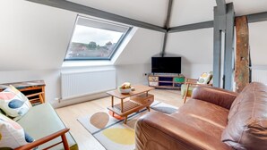 Un salón estilo loft con techo inclinado, tragaluces, televisor, sofá y una mesa pequeña con un frutero.