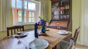 Une salle à manger avec une table en bois dressée pour deux personnes, une bibliothèque et une fenêtre avec des rideaux.