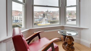 Quarto nos fundos com uma poltrona de couro vermelha, uma mesa de vidro e vista para a paisagem urbana através de amplas janelas.