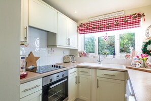 A kitchen with white cabinets, a black oven, a window with a checkered valance, and a sign above the window. - 2 Bed in Holt (oc-2195) (Holt)