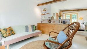 Een gezellige woonkamer met een bank, een houten stoel en een keukenhoek met planken en een raam.