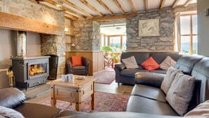 Ein gemütliches Wohnzimmer mit Steinwand, Holzbalken, Kamin, Ledermöbeln und Blick durch große Fenster.