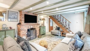 A cozy living room with a brick fireplace, a flat-screen TV, a green cabinet, and a staircase leading to another room. - 3 Bed in Sedgeford (oc-s32725) (Sedgeford)