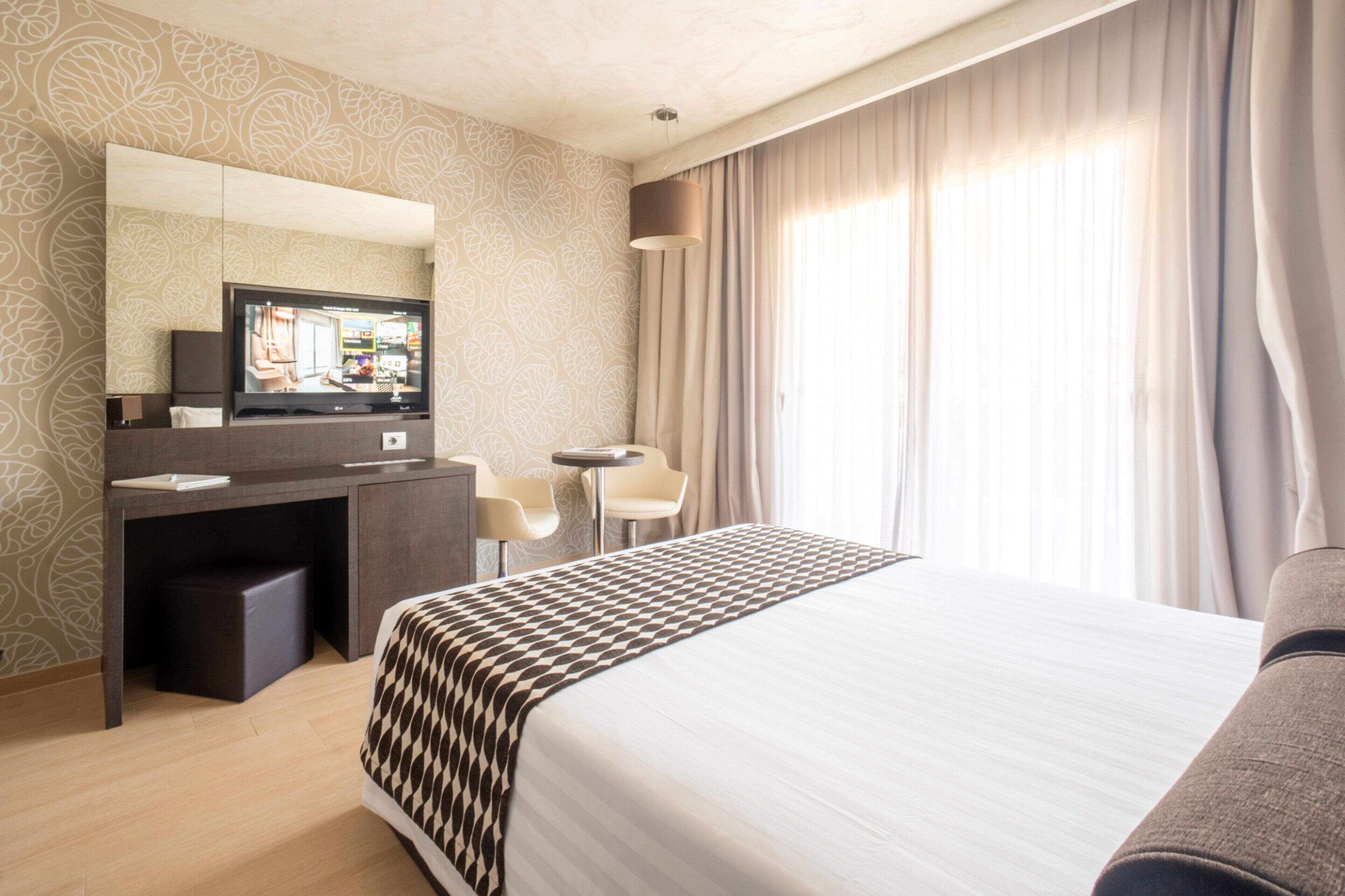A hotel room with a bed, a desk, a chair, a television, and a large window with curtains.