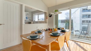 A dining area with a wooden table set for four, surrounded by orange chairs, a hanging light fixture, and a view of a modern building through large windows. - 3 Bed in Saundersfoot (oc-pw522a) (Saundersfoot)