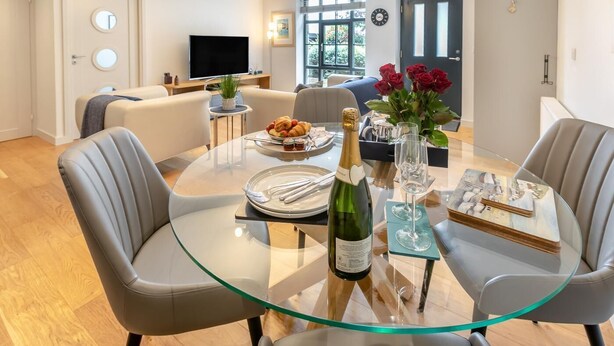 Un comedor moderno con una mesa de vidrio, sillas grises, una botella de champán y un florero con rosas rojas.