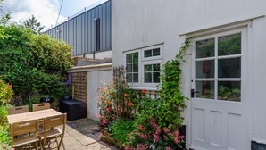 Une petite maison blanche avec une porte et des fenêtres en bois. On y trouve un jardin avec différentes plantes et fleurs, une table en bois et un barbecue.