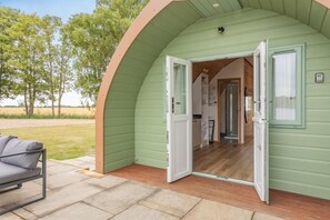 Una cabaña acogedora de una sola habitación con suelo de madera, un pequeño rincón de cocina y una puerta que da a un porche con una silla.