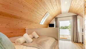 A cozy bedroom with a wooden ceiling, a bed with beige bedding and rolled towels, a TV on a stand, and a view of a lake through a glass door.