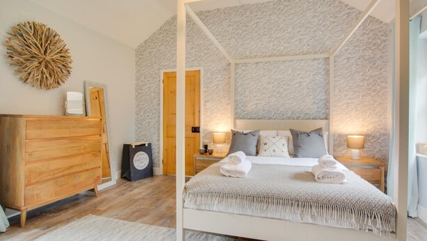 Una cama con dosel de estructura blanca y una pared texturizada al fondo. A la izquierda, un aparador de madera y, a la derecha, una mesita con una lámpara.