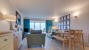 Een moderne woonkamer met een bank, een eettafel met stoelen, een televisie en een groot raam met gordijnen.
