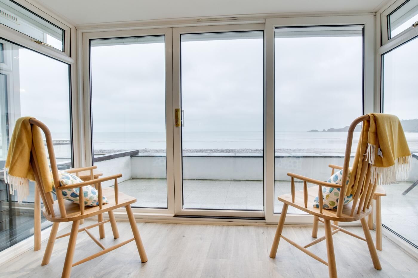 Une pièce avec deux chaises en bois et une porte vitrée donnant sur un balcon avec vue sur la mer.
