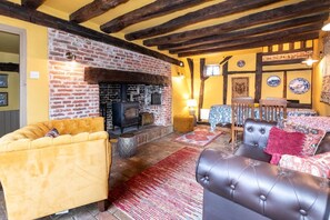 Un salon chaleureux avec une cheminée en briques, des poutres apparentes, un canapé jaune, un fauteuil en cuir et un tapis à motifs.