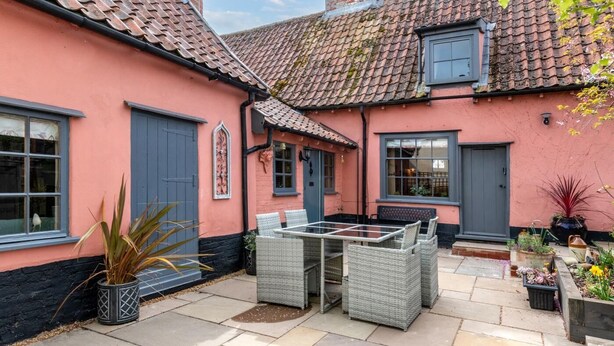 Un bâtiment rose avec un toit en tuiles, une porte bleue et une terrasse meublée de meubles en osier.