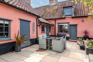 Un bâtiment rose avec un toit en tuiles, une porte bleue et une terrasse meublée de meubles en osier.