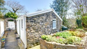 Un bâtiment en pierre doté d’une petite fenêtre, d’un espace extérieur abrité avec des sièges et d’un jardin où poussent différentes espèces de plantes.
