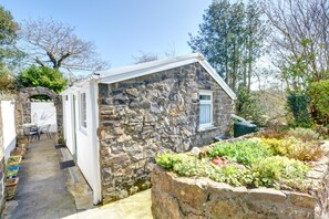 Un bâtiment en pierre doté d’une petite fenêtre, d’un espace extérieur abrité avec des sièges et d’un jardin où poussent différentes espèces de plantes.