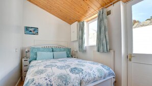 A bedroom with a wooden ceiling, a bed with floral bedding, a nightstand, a window with curtains, and a radiator. - 2 Bed in Saundersfoot (oc-pw907a) (Saundersfoot)