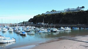 Un port de plaisance où sont amarrés de nombreux bateaux, une plage de sable et, au loin, un grand bâtiment perché sur une falaise.