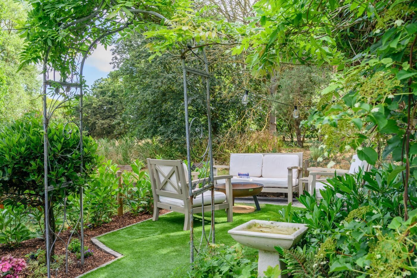 A garden with a white outdoor seating area, including a sofa and chairs, surrounded by lush greenery and a small birdbath.