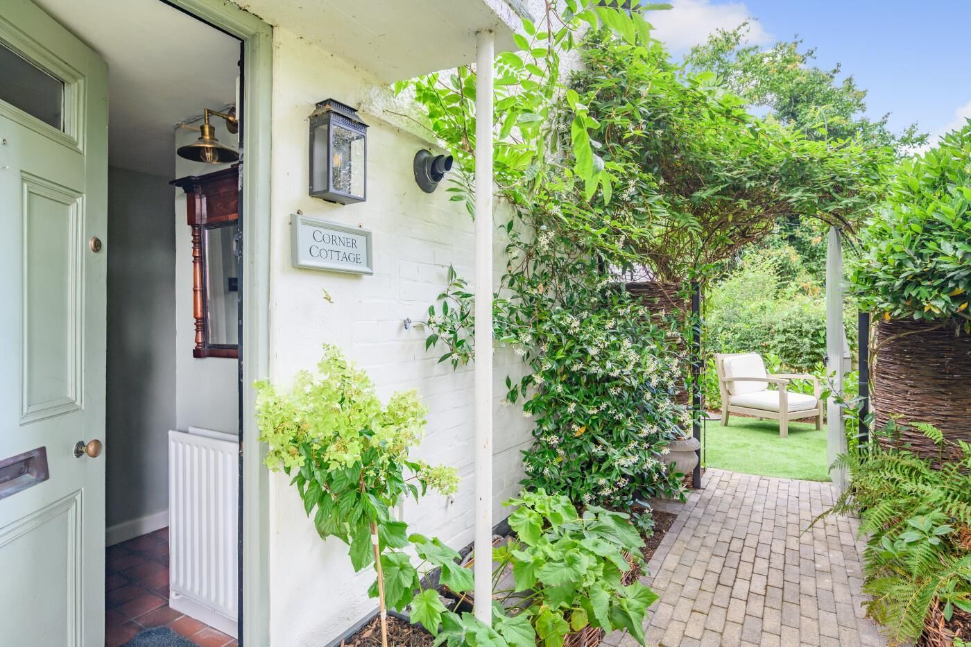 A white cottage with a sign reading "Corner Cottage," a wooden door, a brick pathway, and lush greenery.