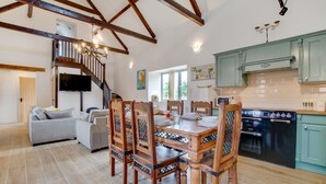 A spacious living room with a wooden dining table and chairs, a staircase, and a kitchen with teal cabinets and a black oven. - 3 Bed in Shereford (oc-2173) (Shereford)