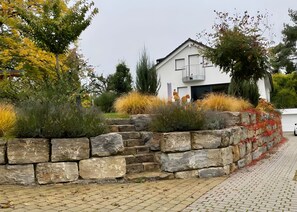 A modern house with a stone retaining wall, a paved driveway, and a variety of plants.