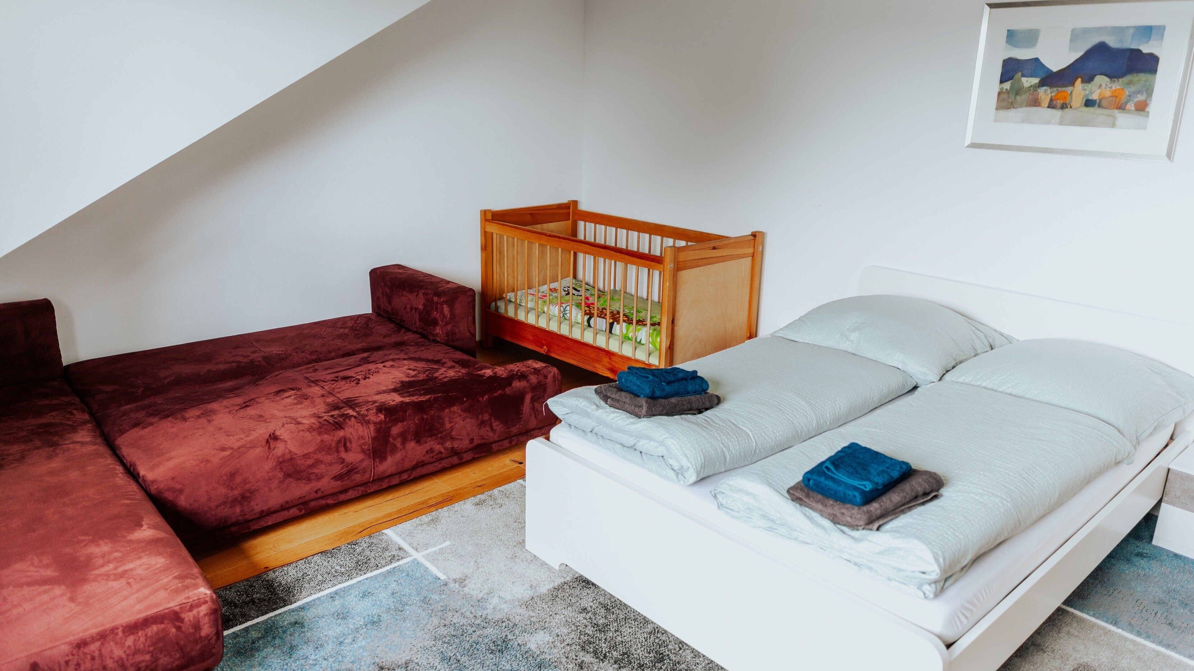 Ein Zimmer mit einem roten Sofa, einem weißen Bett, einem Holz-Babybett und einem Bild an der Wand.