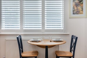 A dining area with a round table, two chairs, and a window with blinds. - 1 Bed in Yapton (oc-b32757) (Yapton)