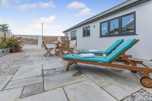 A patio with stone flooring, wooden lounge chairs, a table with chairs, and a white building with windows. - 4 Bed in Padstow (oc-r32776) (Padstow)