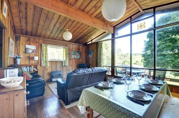 A cozy cabin interior with a dining table set for four, a leather sofa, wooden walls, and large windows offering a view of the outdoors.