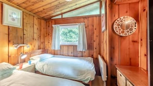 A wooden cabin room with two beds, a window with curtains, a wall-mounted lamp, and a decorative metal plate. - 3 Bed in Broad Haven (oc-wav604) (Haverfordwest)