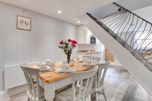 Un coin repas avec une table en bois dressée pour quatre personnes, des chaises blanches, un vase de fleurs et, au fond, une cuisine.