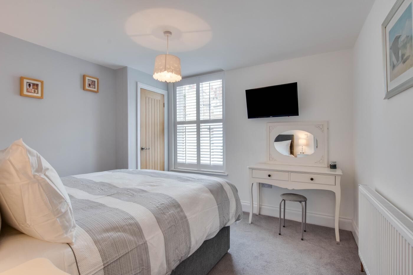 Een slaapkamer met een bed, een bureau met spiegel, een televisie en een raam met jaloezieën.