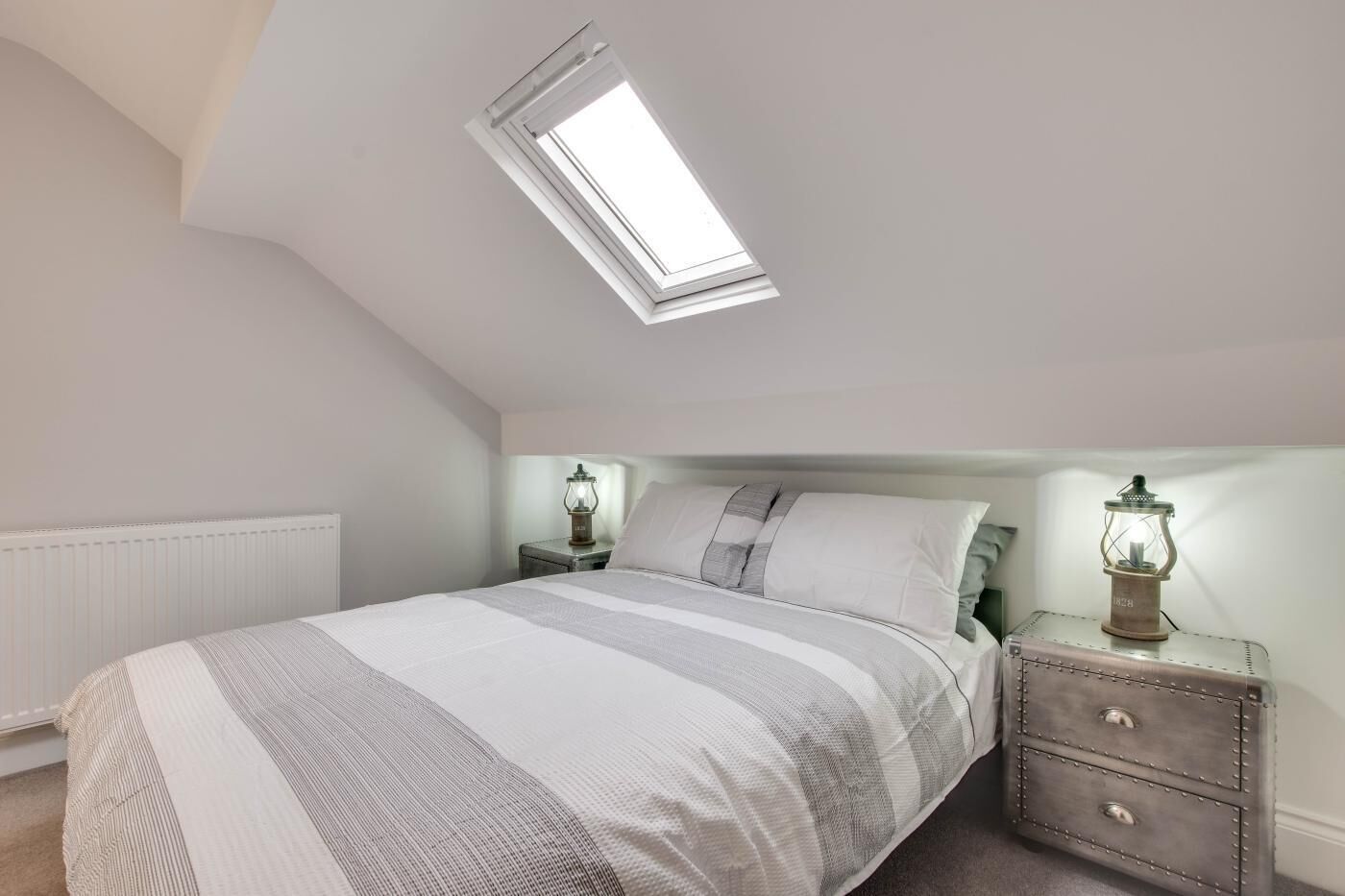 A bedroom with a skylight, a bed with striped bedding, two bedside tables with lantern-style lamps, and a radiator.