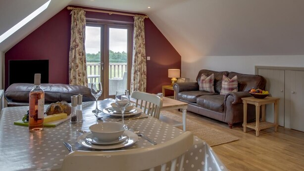 A dining area with a table set for two, a leather sofa, and a window with curtains. - 1 Bed in Swannington (oc-2206) (Attlebridge)