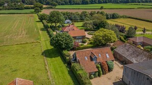 An aerial view of a rural property with multiple buildings, expansive green fields, and a forested area in the background. - 1 Bed in Swannington (oc-2206) (Attlebridge)