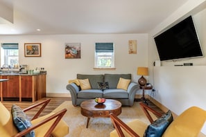 A living room with a sofa, coffee table, and chairs, a kitchen area with a sink and counter, and a TV mounted on the wall. - 3 Bed in Birnam (oc-o32901) (Birnam)