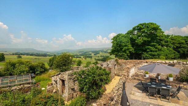 Una estructura de piedra con una mesa y sillas, con vistas a un paisaje verde y colinas distantes.