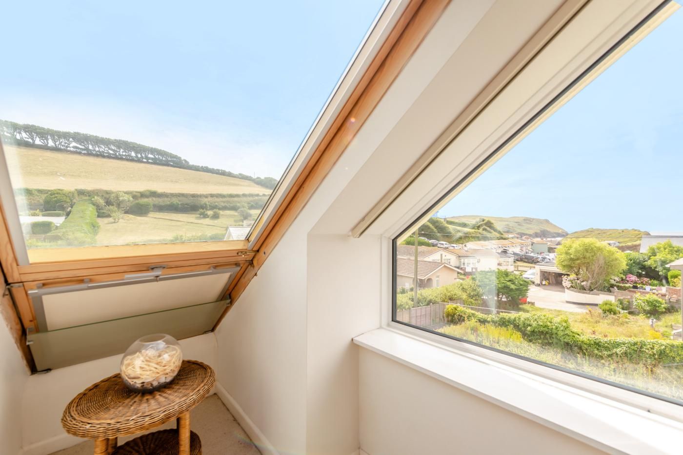 A skylight window with a view of a rural landscape and a small wicker table with a bowl.