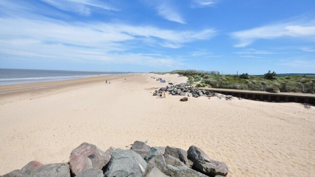 A wide sandy beach with a wooden boardwalk, rocky outcrops, and sparse vegetation. - 3 Bed in Brancaster (oc-2153) (Brancaster)
