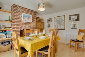 A dining area with a brick fireplace, a yellow tablecloth, and wooden chairs. - 3 Bed in Brancaster (oc-2153) (Brancaster)