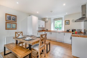 A modern kitchen with white cabinets, a wooden dining table, and a window providing natural light. - 2 Bed in Crooklands (oc-c32881) (Crooklands)