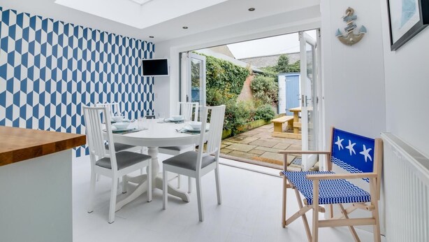 A modern kitchen with a dining area, a TV mounted on the wall, and a view of the garden through the glass door. - 2 Bed in Southwold (oc-19cs) (Southwold)