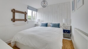 A bedroom with a large bed, a wooden-framed mirror, a window with blinds, a bedside table with a lamp, and a radiator. - 2 Bed in Southwold (oc-19cs) (Southwold)