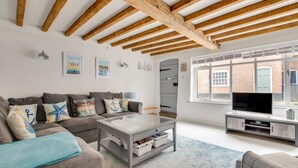A living room with a sofa, coffee table, TV, and a window with a view of a brick building. - 2 Bed in Southwold (oc-19cs) (Southwold)