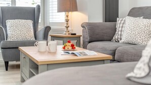 Ein gemütliches Wohnzimmer mit einem grauen Sofa, einem passenden Sessel und einem Holztisch mit einem Buch und einer Auswahl an Gebäck.