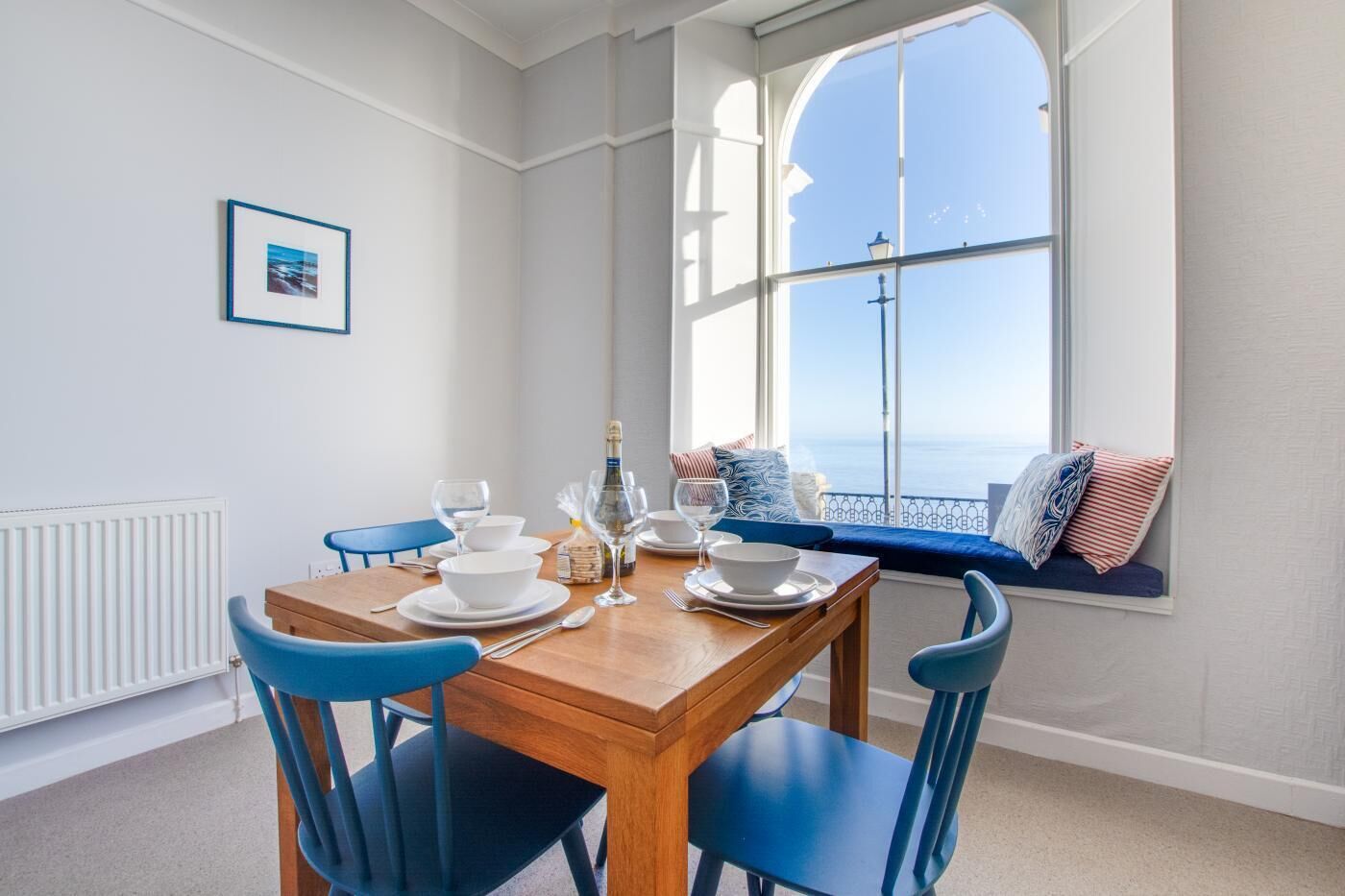 Un coin repas avec une table en bois dressée pour quatre personnes, des chaises bleues et une vue sur la mer par une grande fenêtre.