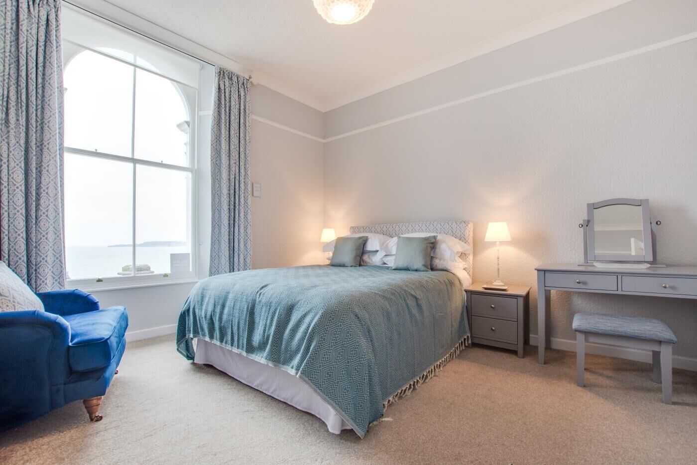 A bedroom with a bed, bedside table, chair, and a desk with a mirror.